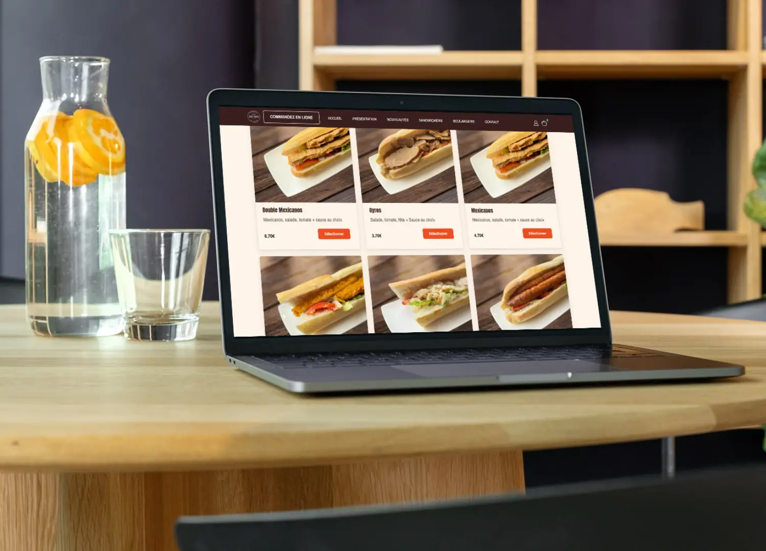 Photo d'une table avec une carafe d'eau avec des rondelles d'oranges, un verre vide et un ordinateur sur la page d'un site web e-commerce réalisé par clickonweb prêt à commander des sandwiches.